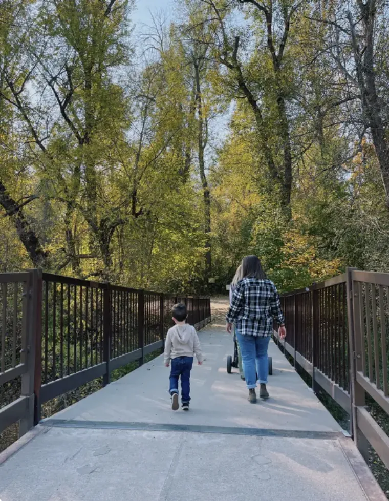 a mom and her young son walking across a bridge into a peaceful wooded area