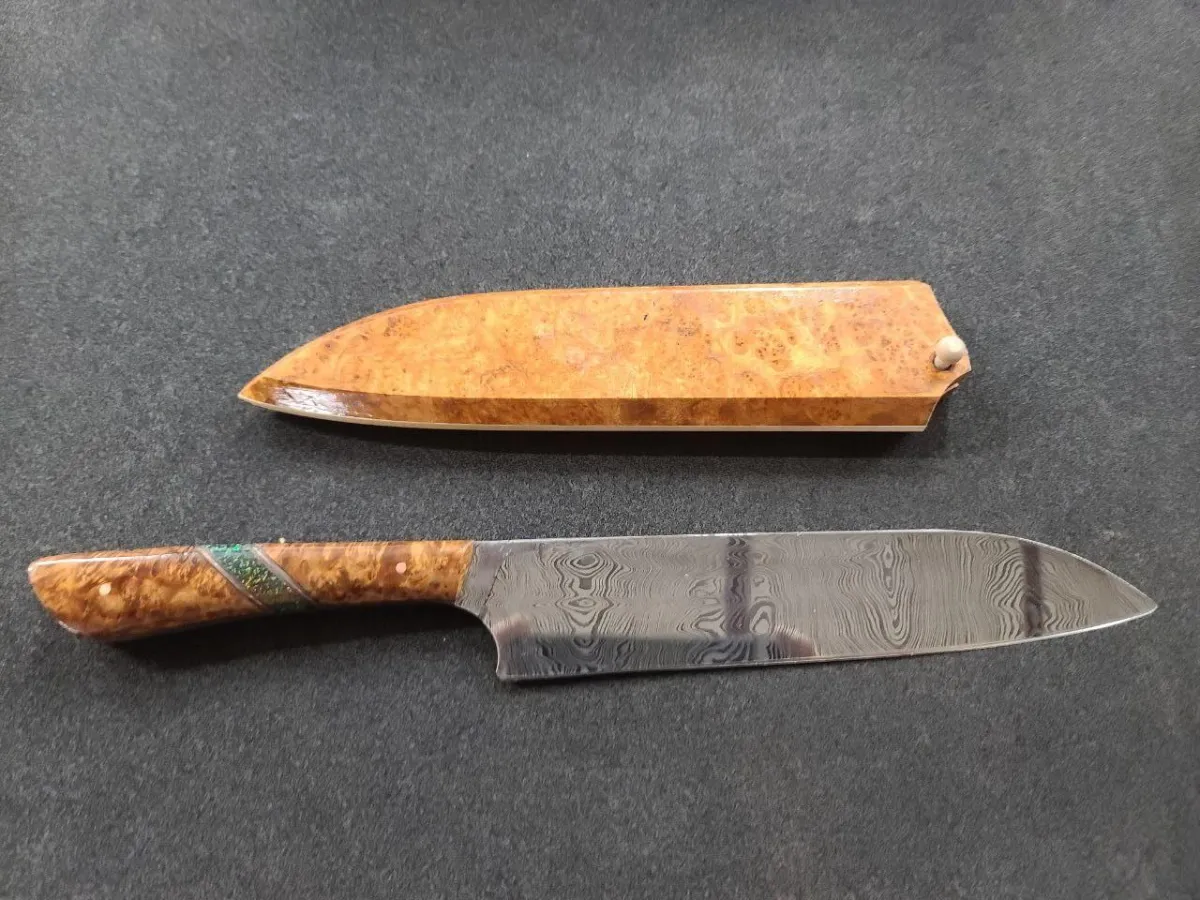 Handcrafted 9-inch Damascus steel Santoku knife with Golden Amboyna burl handle featuring Black Opal and copper spacers. Shown with matching saya (wooden sheath). The blade displays intricate Damascus pattern with pronounced wood grain effect, complemented by the striking figure of the burl wood handle