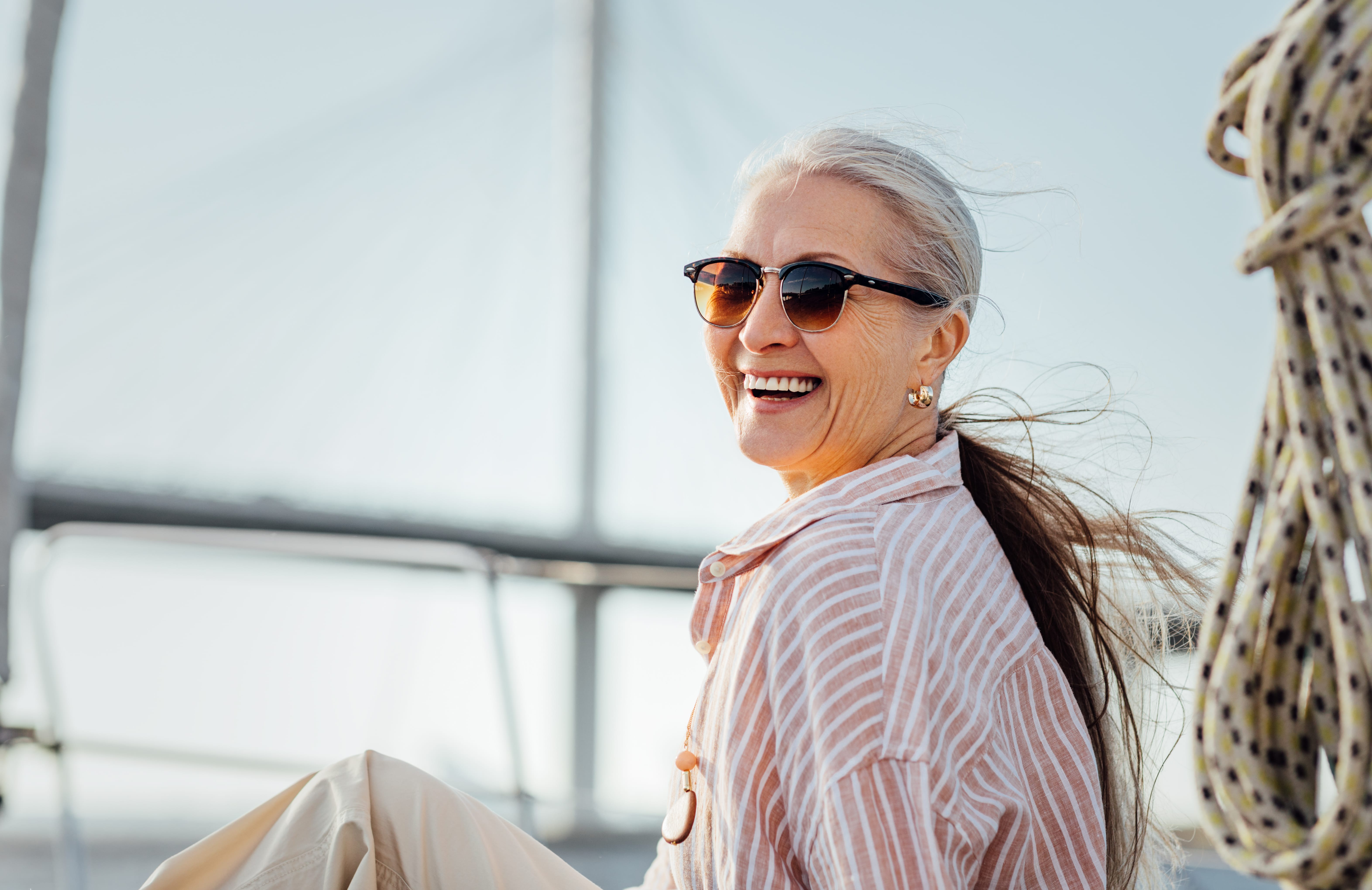 Smiling elderly woman with sunglasses and gray hair in a ponytail, wearing a striped shirt on a sunny day, with a bridge in the background.
