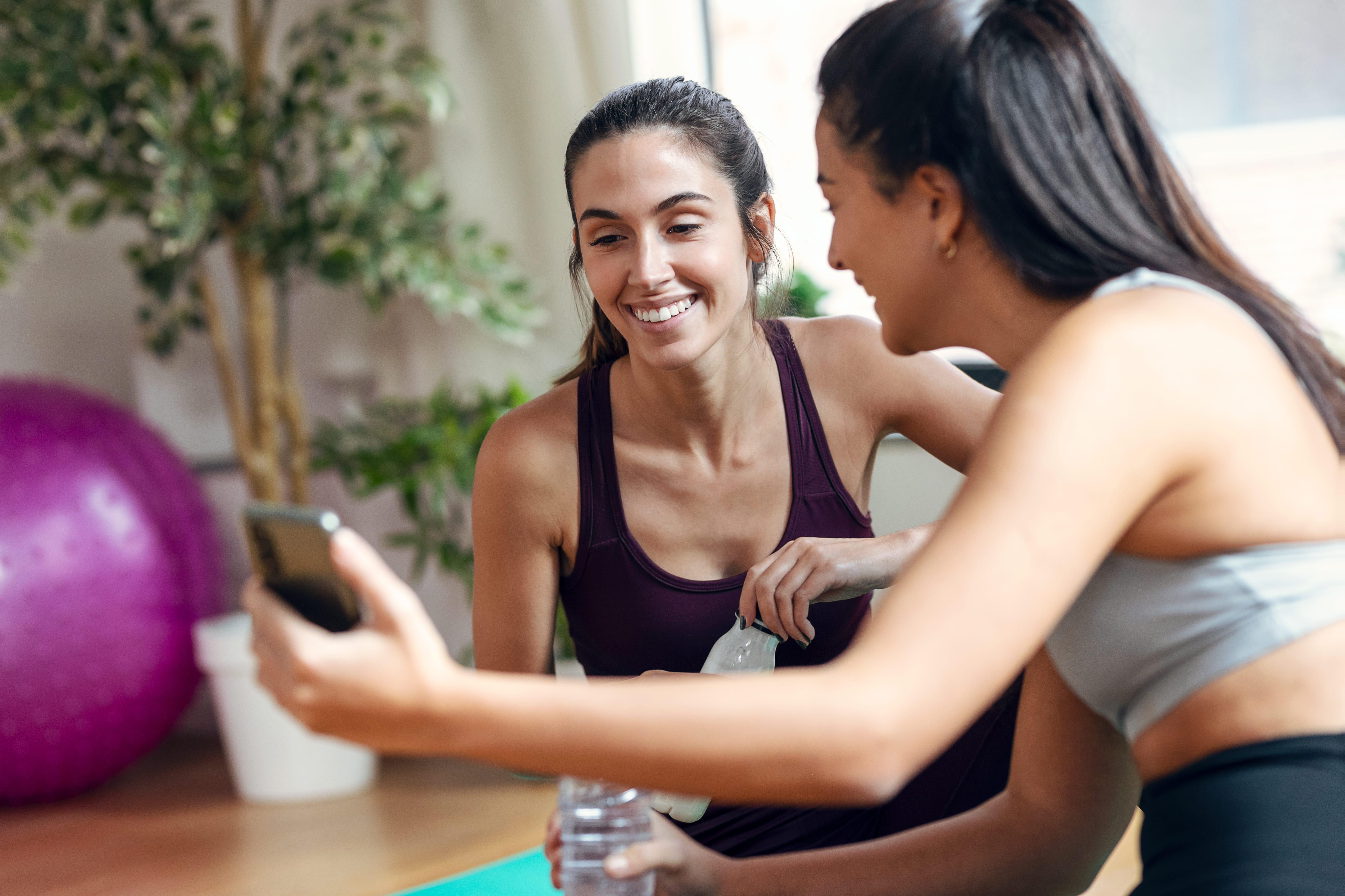Two women in workout gear smile as they look at a smartphone. They hold water bottles, sitting on yoga mats with an exercise ball in the background.