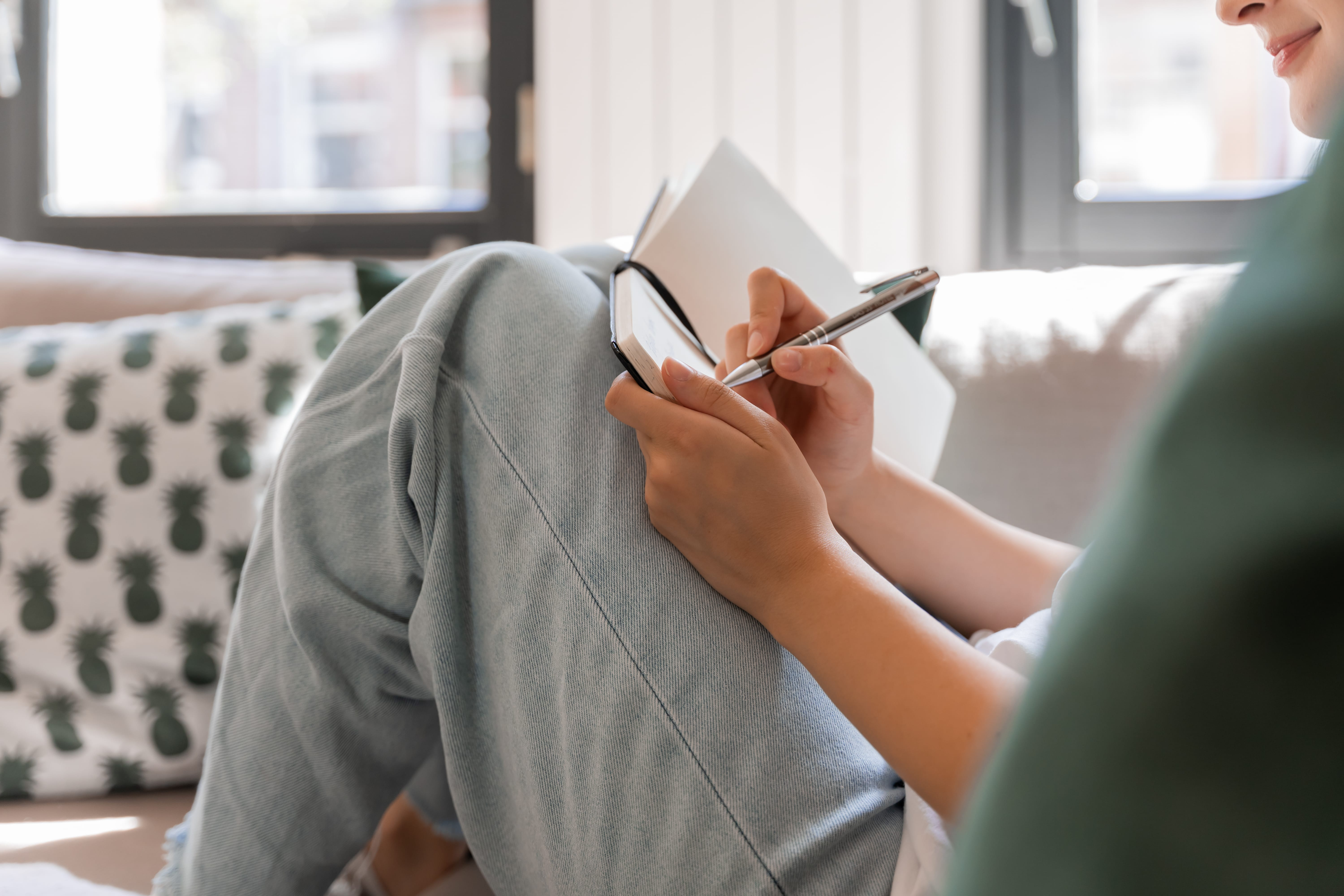 A person sits on a couch with crossed legs, journaling in a notebook. They wear light jeans, and a pineapple-patterned pillow is beside them, in a sunlit room.