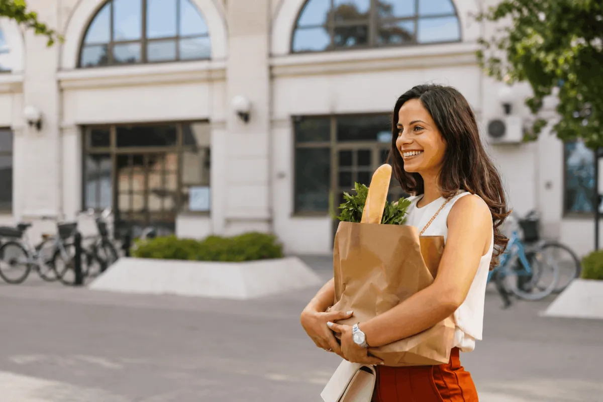 A woman smiles while holding a paper bag filled with fresh vegetables, showcasing her healthy grocery choices.