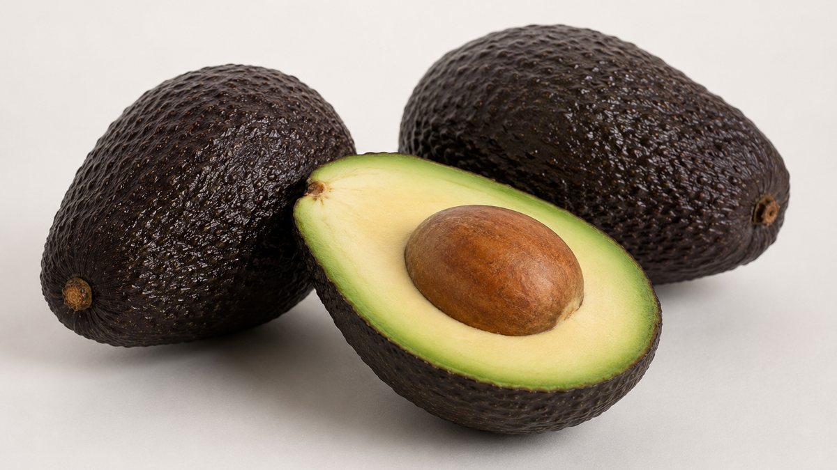 Three ripe Ugandan Hass avocados with dark pebbly skin, one halved to show the buttery yellow-green flesh and intact seed of ripening-programme fruit