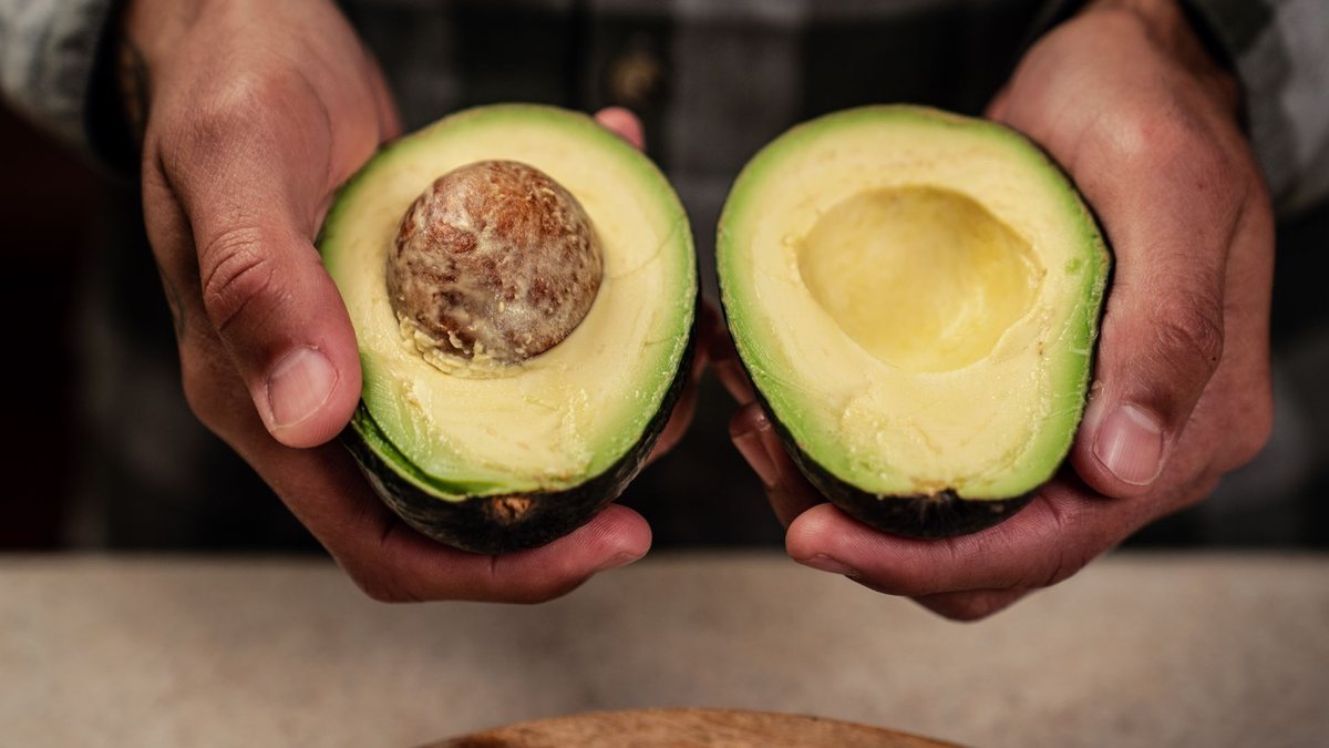 Hands holding a halved Hass avocado with clean pale-green flesh and an intact seed, the even interior that proves maturity at pick
