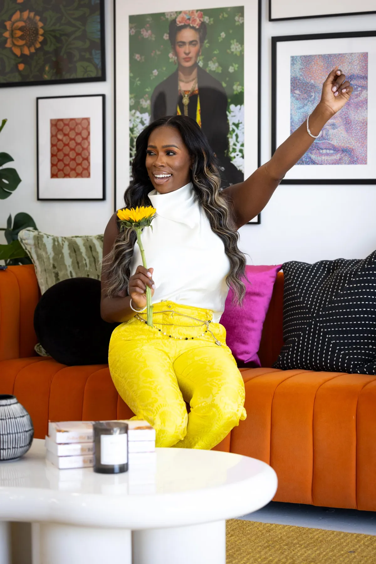 Joyful Black woman , Quellyn Rosa Kennedy , sitting on a vibrant orange couch, holding a sunflower, dressed in bright yellow pants and a white top, exuding confidence and positivity in a stylish, art-filled space.