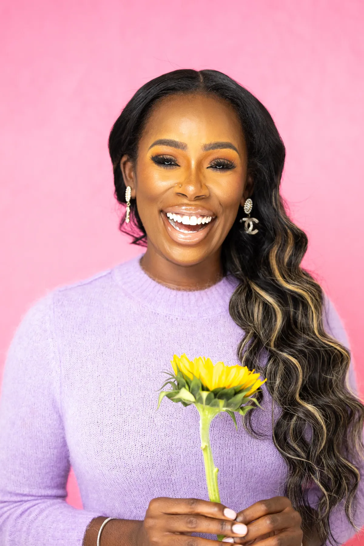 Quellyn Rosa Kennedy, black woman smiling radiantly against a pink background, wearing a lavender sweater and holding a sunflower, exuding warmth, confidence, and joy.