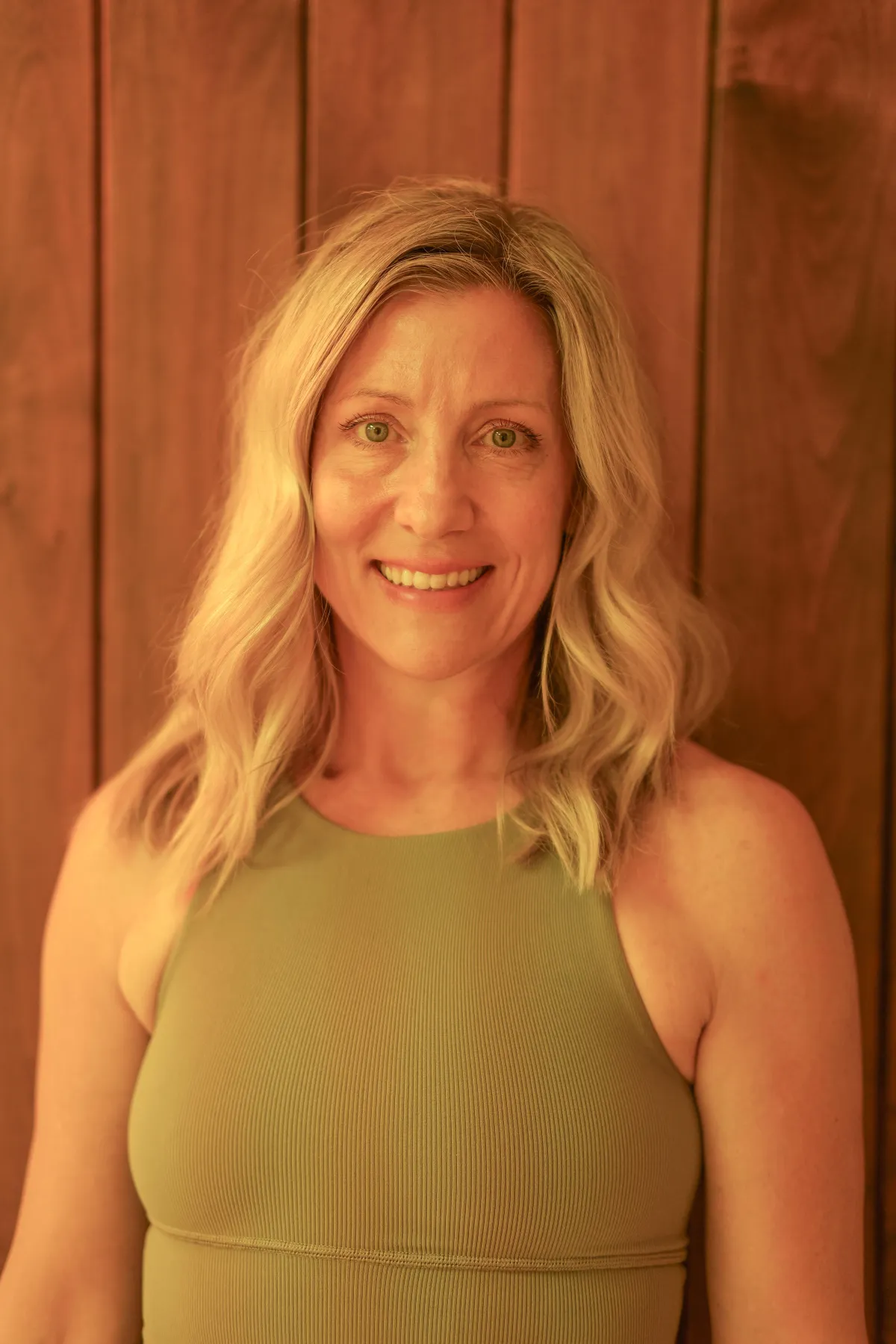 Smiling female yoga teacher at Plymouth Yoga Room wearing a light beige sweatshirt, standing in front of a wooden wall, representing a nurturing, approachable presence dedicated to mindfulness, movement, and holistic wellness.