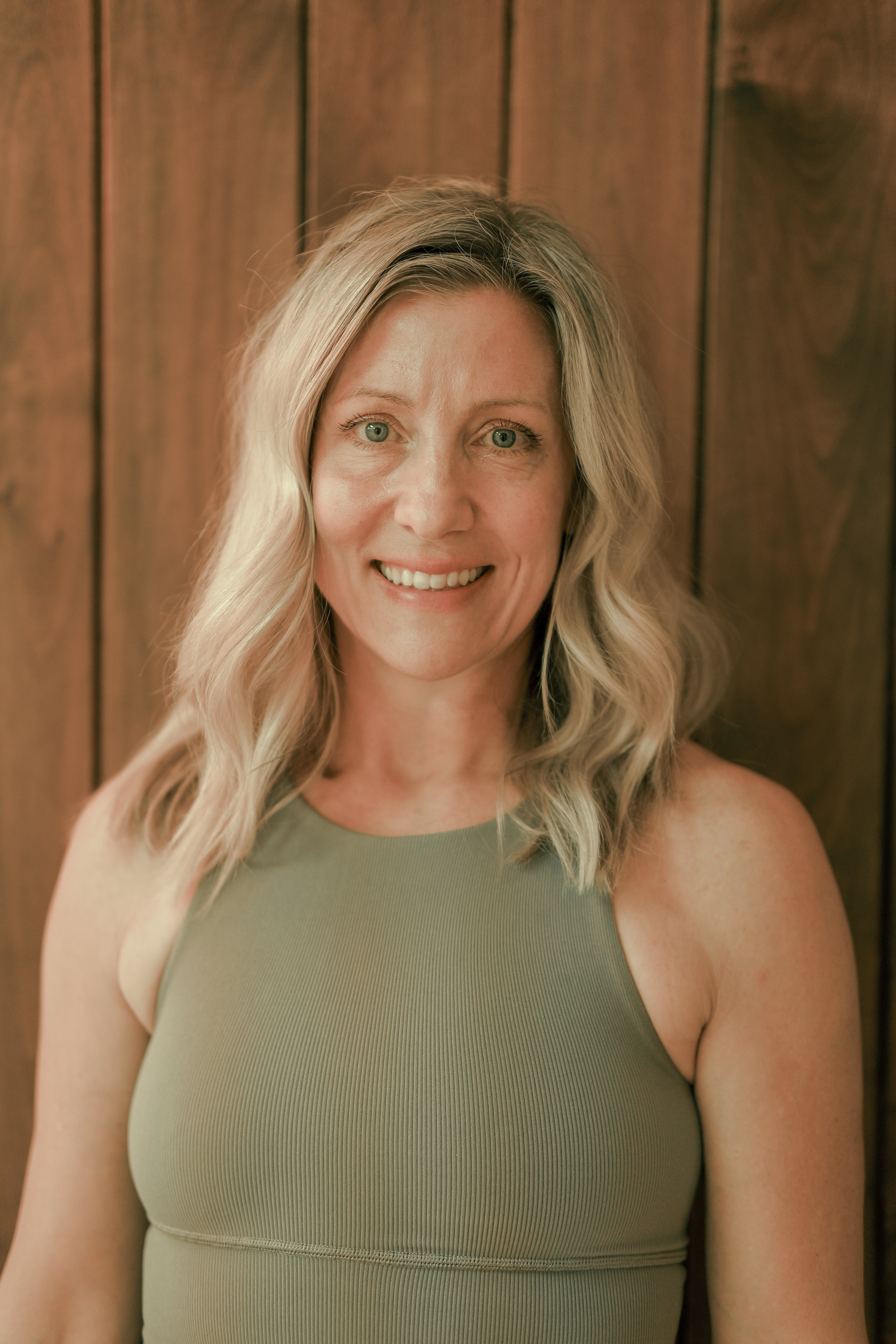 Smiling female yoga teacher at Plymouth Yoga Room wearing a light beige sweatshirt, standing in front of a wooden wall, representing a nurturing, approachable presence dedicated to mindfulness, movement, and holistic wellness.
