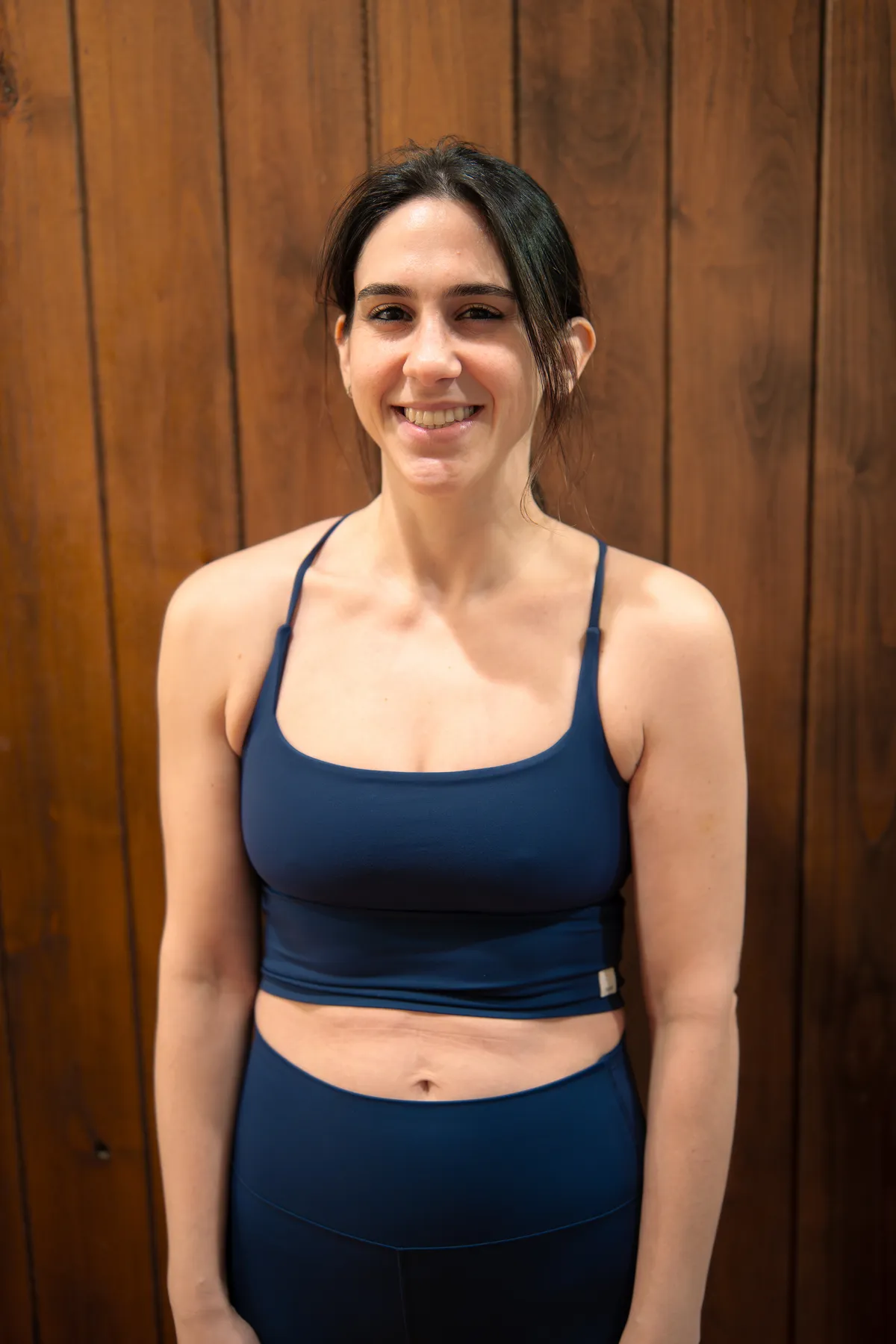 Smiling female yoga teacher at Plymouth Yoga Room wearing a black crop top, standing confidently in front of a wooden wall, representing strength, grace, and a commitment to mindful movement and holistic wellness.
