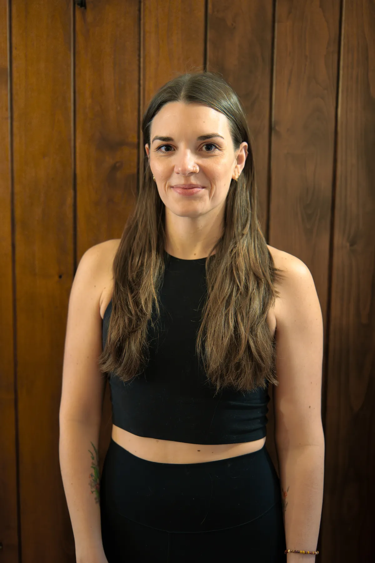 Smiling female yoga teacher at Plymouth Yoga Room wearing a black crop top, standing confidently in front of a wooden wall, representing strength, grace, and a commitment to mindful movement and holistic wellness.