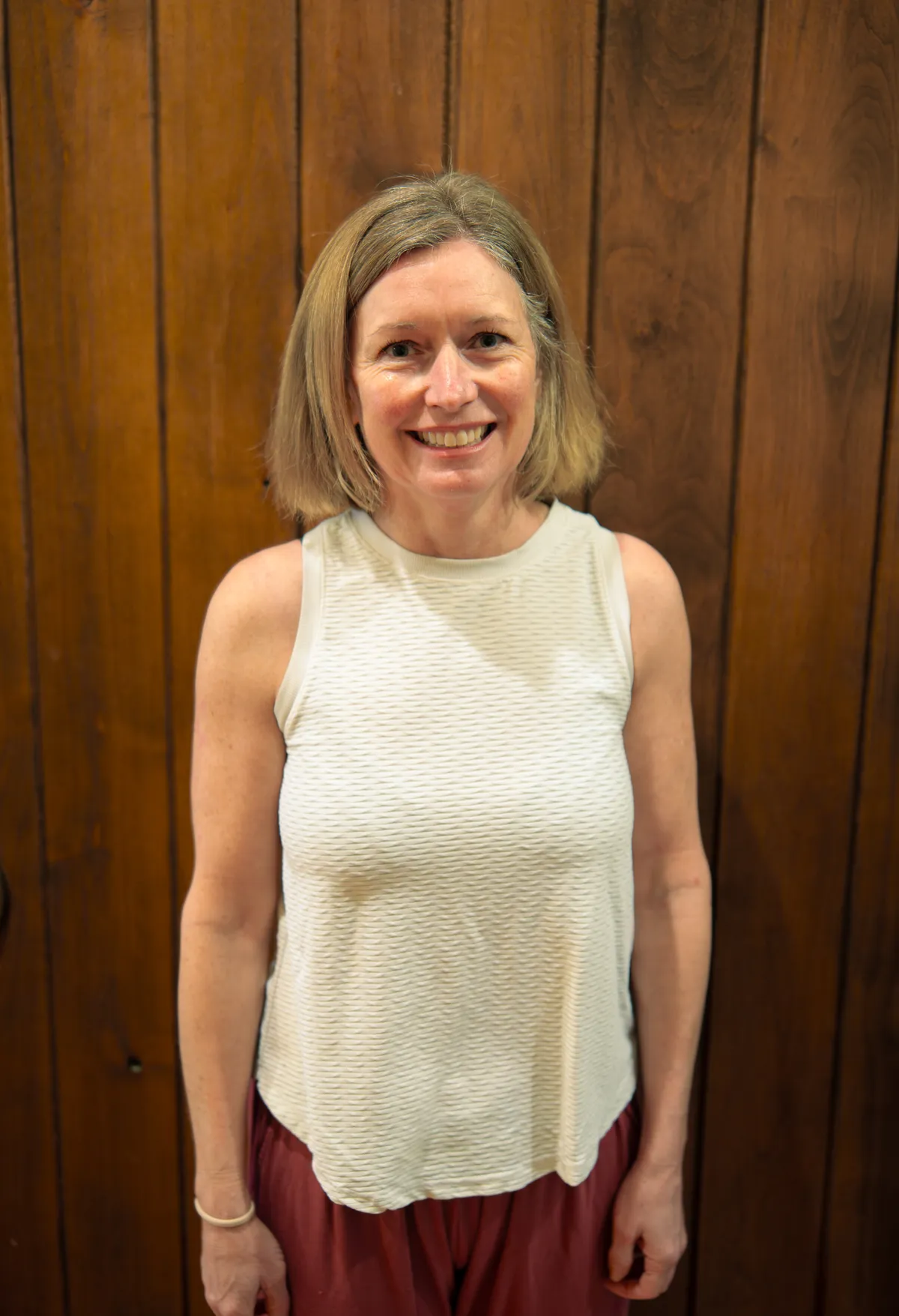 Smiling female yoga teacher at Plymouth Yoga Room wearing a black crop top, standing confidently in front of a wooden wall, representing strength, grace, and a commitment to mindful movement and holistic wellness.