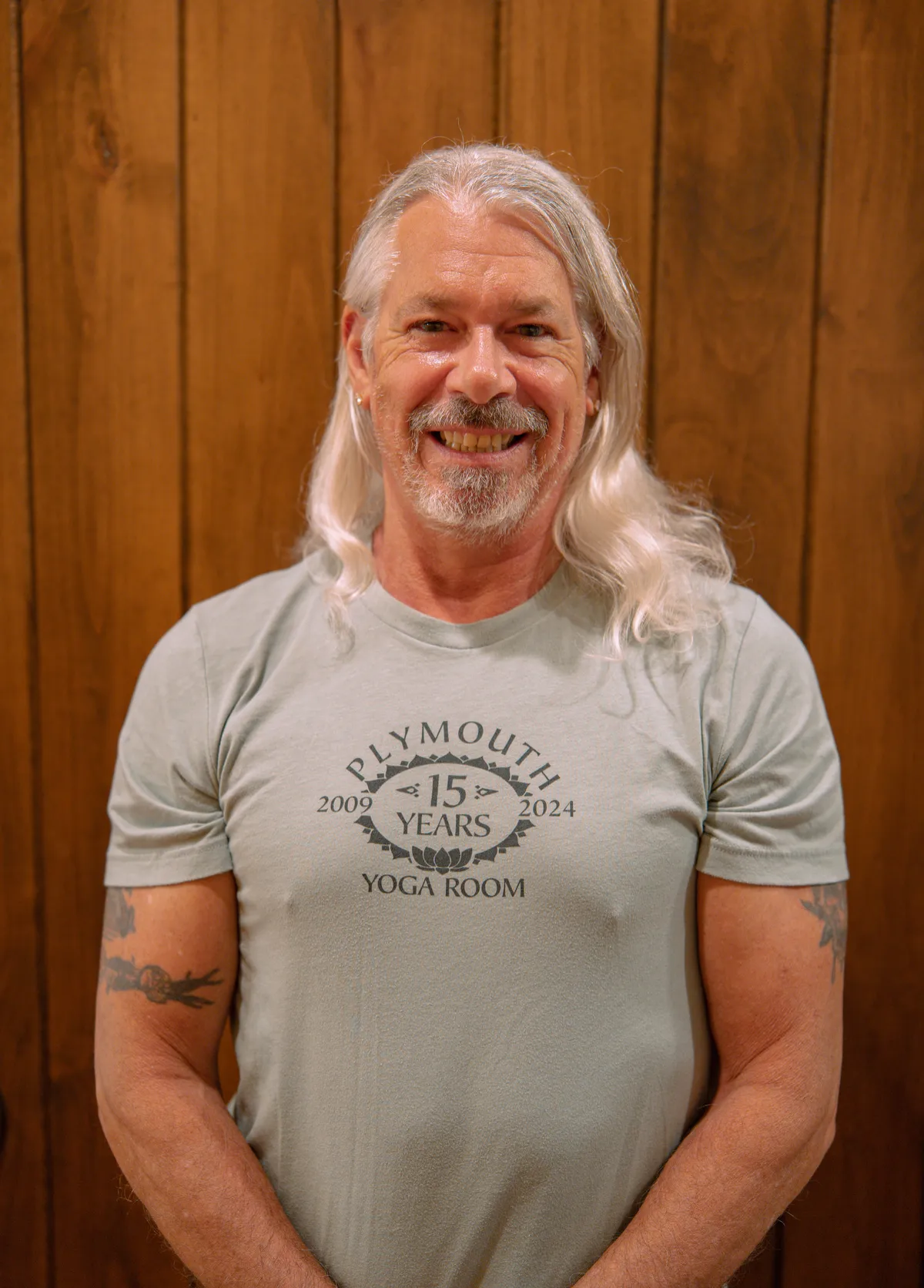 Smiling male yoga teacher at Plymouth Yoga Room wearing a dark "Plymouth Yoga Room Est. 2009" shirt, standing in front of a wooden wall, representing strength, mindfulness, and a welcoming approach to yoga and wellness.