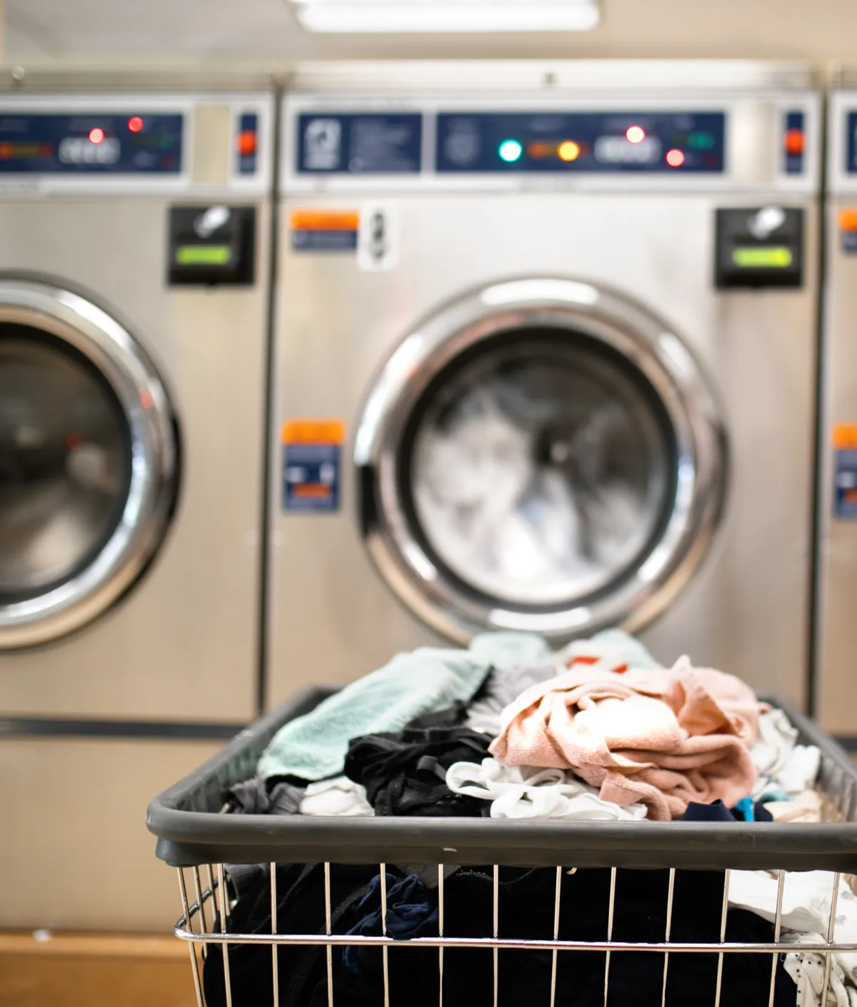 Three laundry machines sit side by side whirring while washing clothes. A hamper of dirty laundry sits in front of them. 