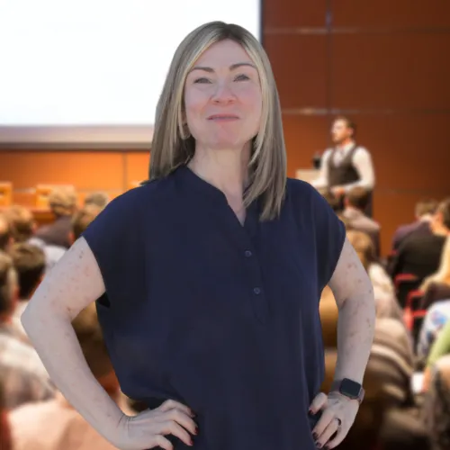 Confident woman with straight blonde hair standing with hands on hips, smiling slightly. She is wearing a dark blouse and a smartwatch, posed in front of a blurred background of a lecture hall with an audience and a speaker on stage.