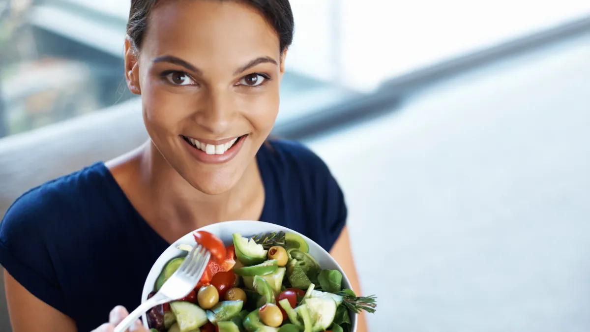 Smiling woman holding a fork and a bowl of fresh salad, looking up at the camera. The salad contains lettuce, tomatoes, cucumbers, olives, and avocado, suggesting a healthy meal choice.