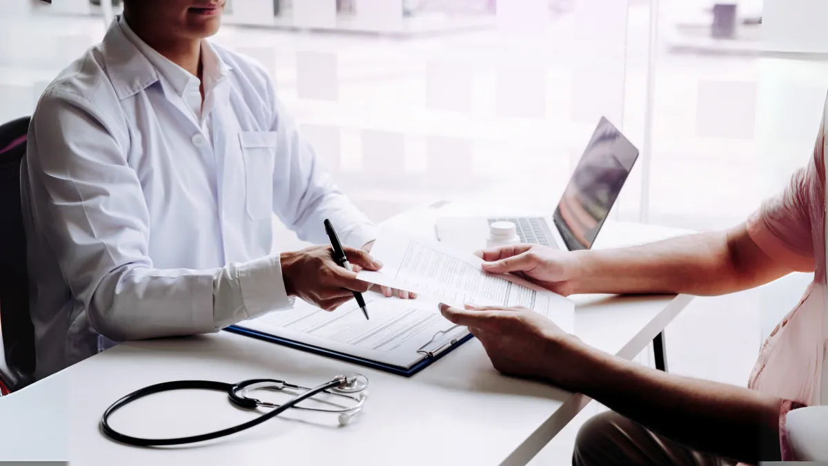 Doctor in a white coat sitting at a desk with a stethoscope and laptop, handing a medical form or prescription to a patient. The scene suggests a professional consultation or health assessment in a clinical setting.