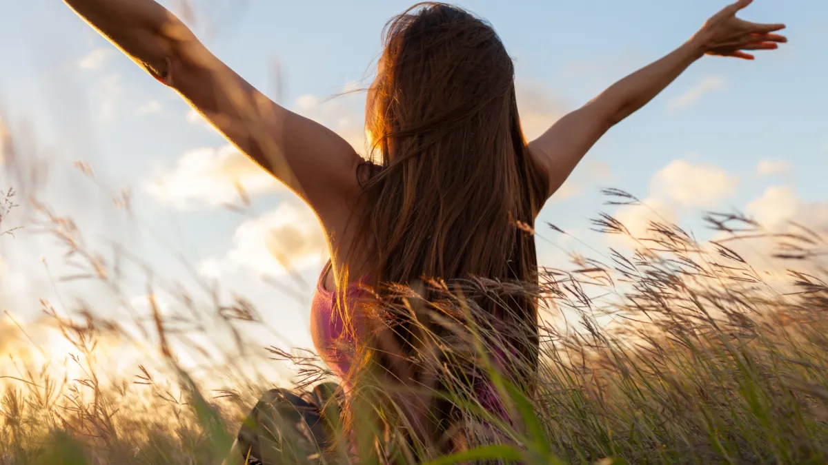Woman sitting in a field of tall grass with her arms raised joyfully toward the sky. The sun is setting in the background, casting a warm golden light, symbolising freedom, peace, and connection with nature.