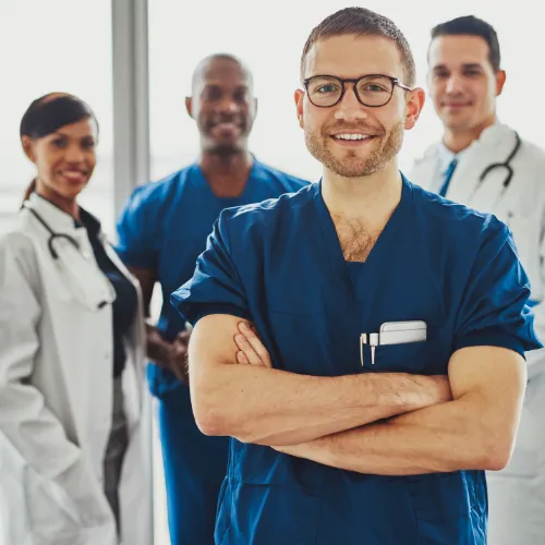 Smiling male healthcare professional in blue scrubs standing confidently with arms crossed, with three other medical team members in the background, all wearing professional attire and stethoscopes, indicating a collaborative and diverse medical team.