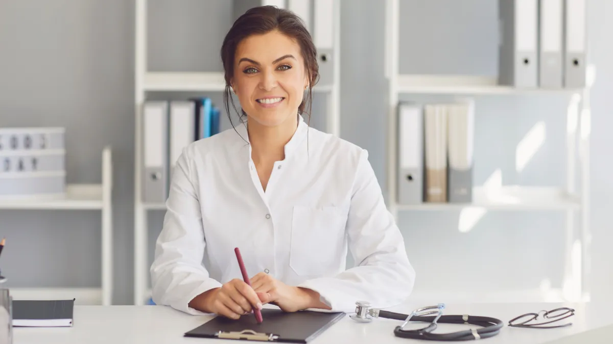 Smiling female doctor sitting at a desk with a clipboard and pen, wearing a white lab coat. A stethoscope and glasses are placed on the desk, and shelves with organized folders are visible in the background, indicating a professional medical office.
