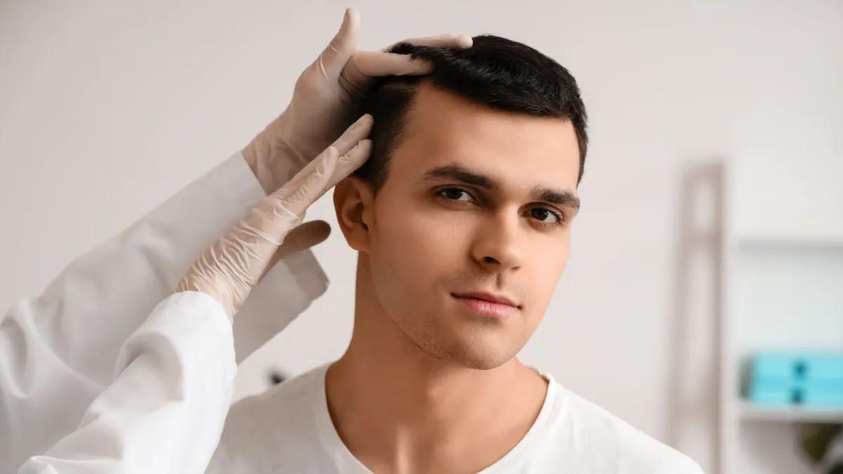 Young man with short dark hair receiving a scalp examination from a medical professional wearing white gloves. The setting appears to be a clinic or consultation room, suggesting a hair or dermatology check-up.