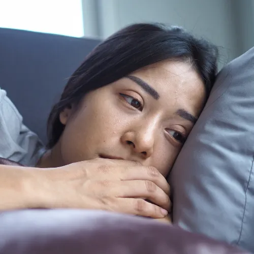 Young woman lying on a couch with her head resting on a pillow, looking sad or deep in thought. Her eyes appear tired and her hand gently touches her face, suggesting feelings of sadness, fatigue, or emotional distress.
