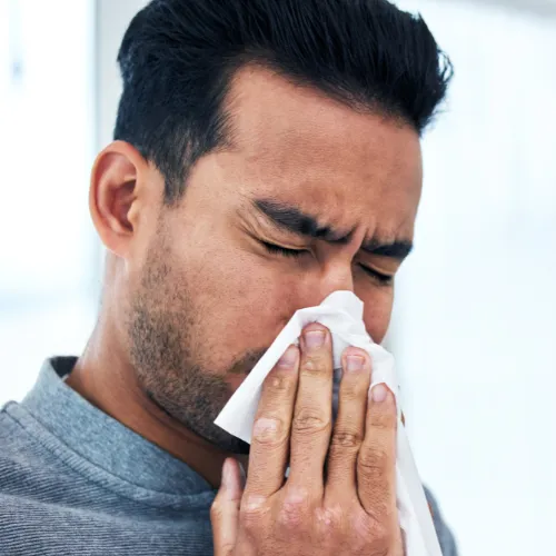 Man holding a tissue to his nose while sneezing or blowing his nose. His eyes are shut tightly, and he appears to be experiencing symptoms of a cold or allergies, with a pained expression on his face.