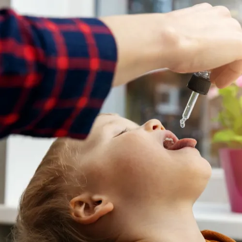Young child with mouth open, receiving liquid medicine or supplement from a dropper held by an adult. The child is indoors, and the background shows a window with soft daylight and a pink flower pot.