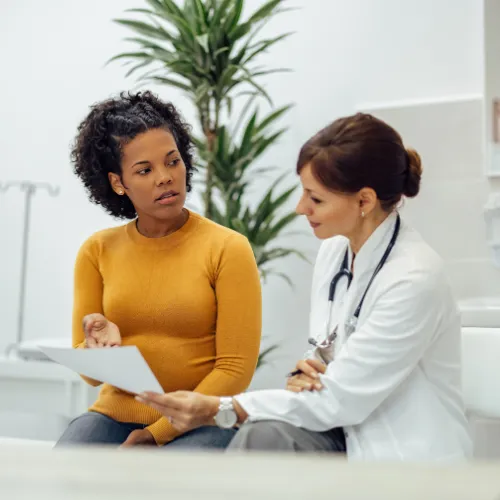 Female doctor in a white coat reviewing medical documents with a female patient wearing a mustard yellow sweater. The two are seated in a clinic setting, engaged in a serious and attentive conversation.