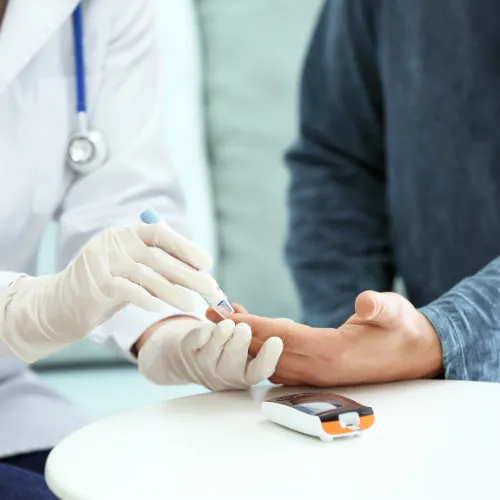 Healthcare professional wearing gloves using a lancet device to prick a patient’s finger for a blood glucose test. A glucometer is placed on the table in front of them, indicating a diabetes or blood sugar monitoring procedure.