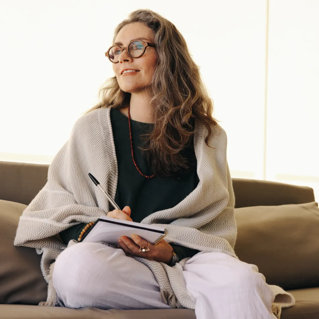 Smiling middle-aged woman with long grey hair and glasses, sitting on a couch with a notepad and pen in hand. She is wrapped in a light shawl and appears thoughtful or reflective, looking slightly upward. The setting suggests a calm, indoor environment with soft natural light.