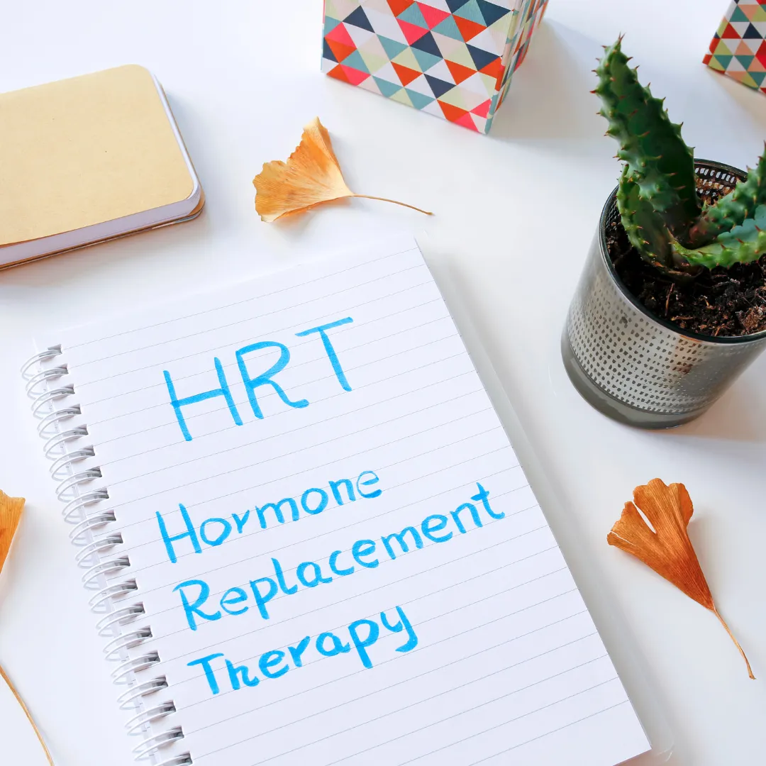 Notebook on a white desk with the handwritten words ‘HRT Hormone Replacement Therapy’ in blue ink. Surrounding the notebook are dried ginkgo leaves, a small potted cactus, a closed notepad, and a colourful geometric-patterned box, creating a bright and tidy workspace.