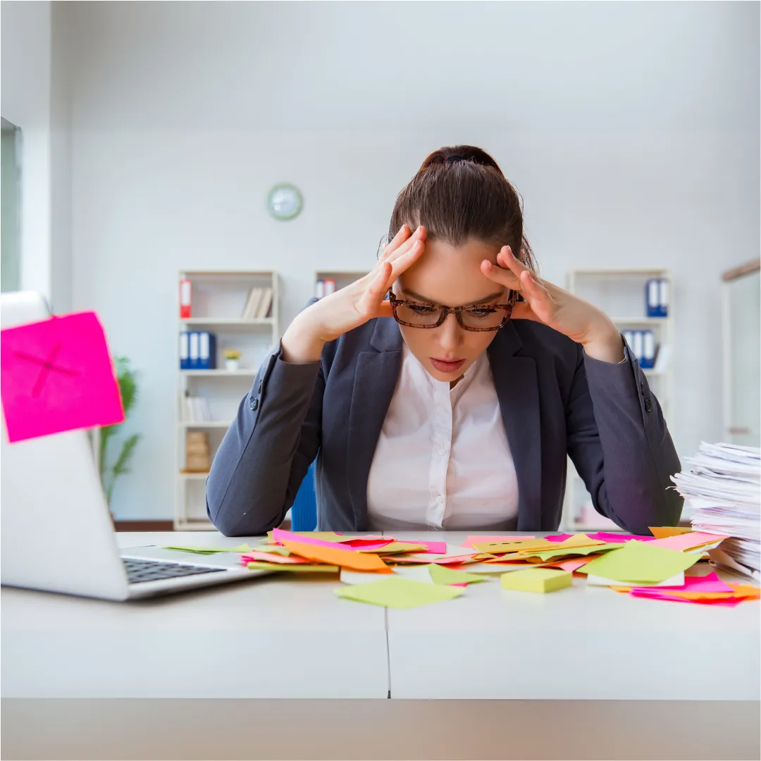 Stressed businesswoman sitting at a cluttered desk covered in colourful sticky notes and papers. She is holding her head with both hands and looking down in frustration. A laptop and a large stack of documents are also visible, indicating overwhelm or workplace burnout.