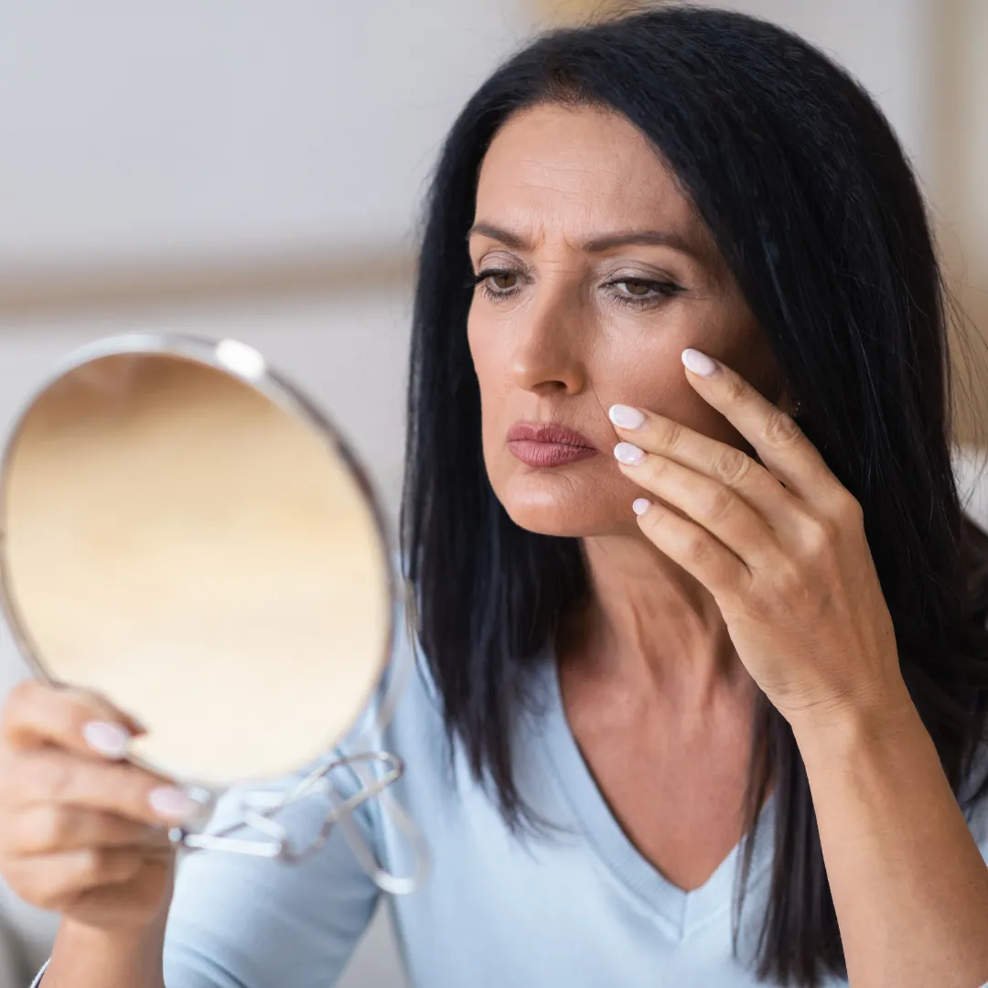 Middle-aged woman with long dark hair examining her face closely in a handheld mirror. She appears concerned or focused as she gently touches the skin on her cheek, possibly checking for signs of ageing or skin issues.