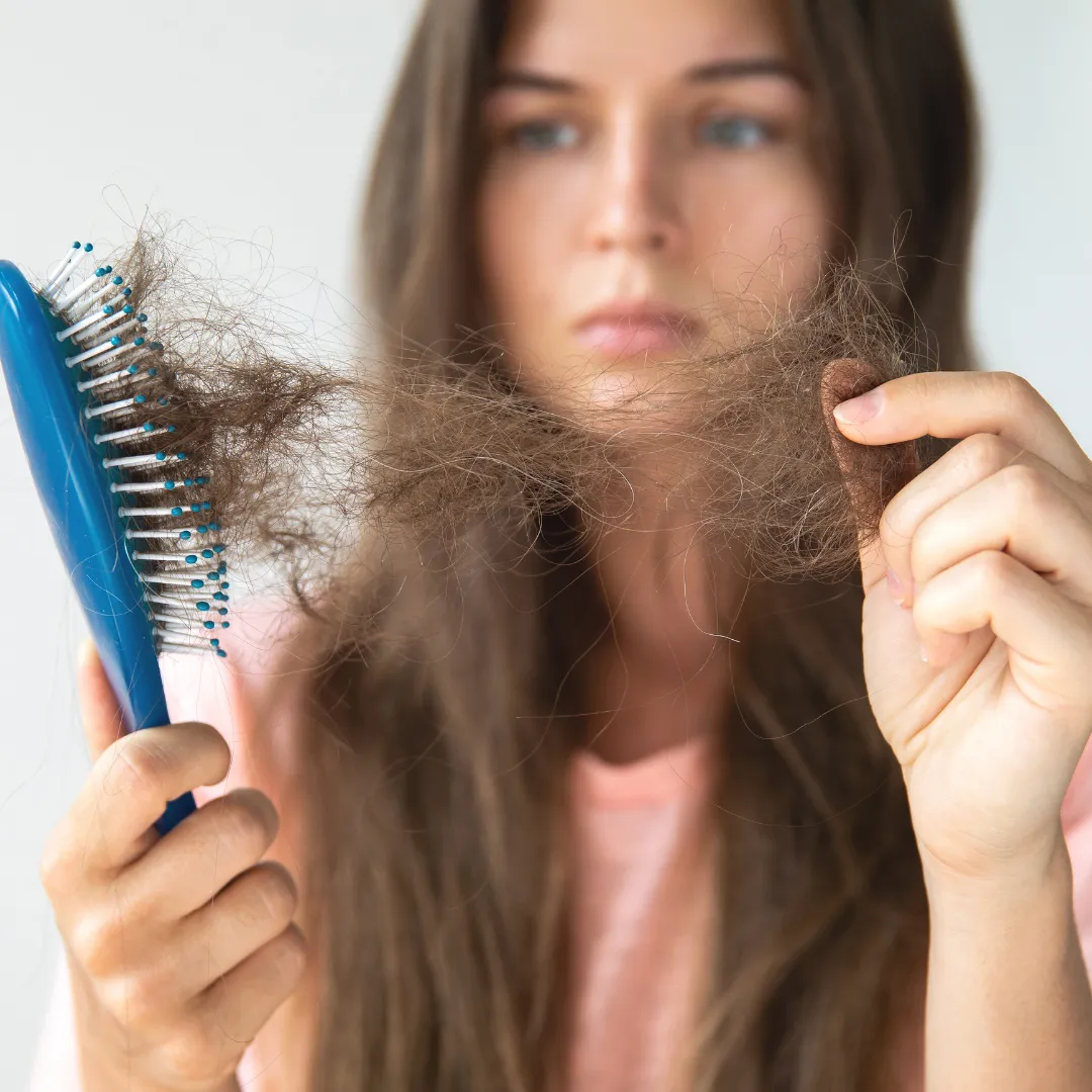 Woman with long brown hair looking concerned while holding a hairbrush full of tangled, fallen hair in one hand and loose strands in the other, indicating hair loss or excessive shedding.