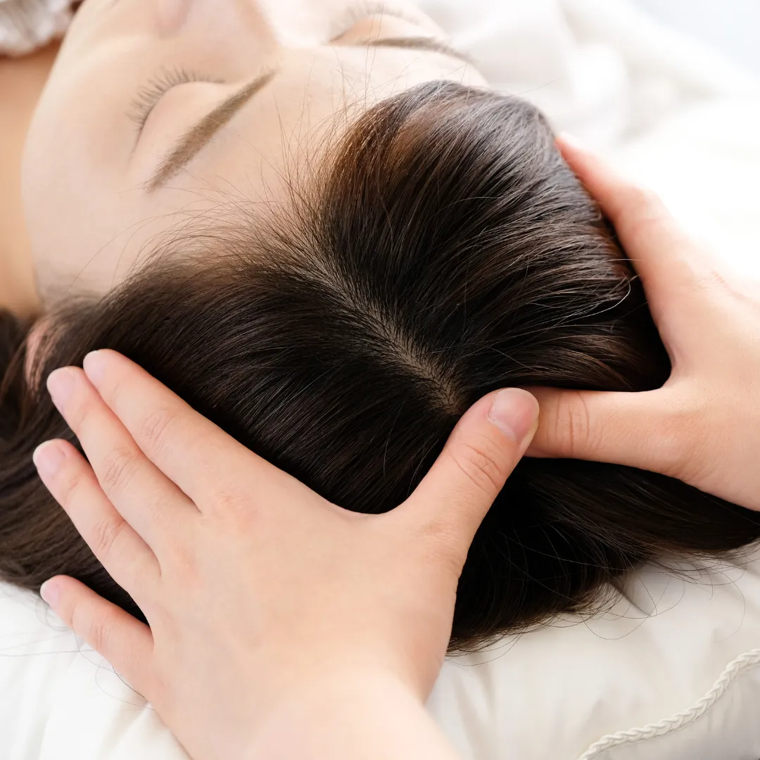 Close-up of a woman lying down with eyes closed while receiving a gentle scalp massage. Hands are applying pressure to her head, suggesting a relaxing or therapeutic hair and scalp treatment.