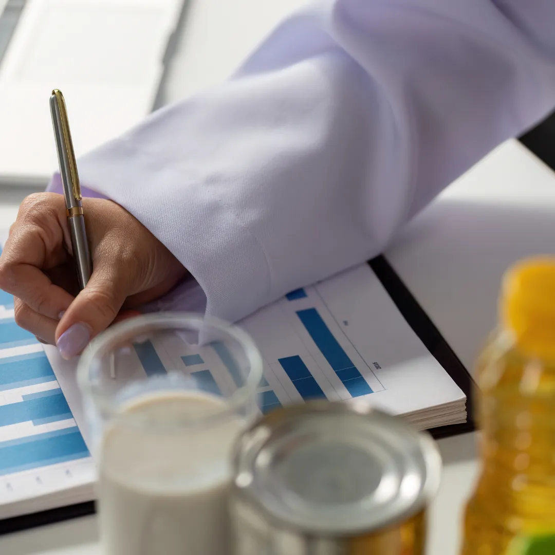 Close-up of a person in a white lab coat holding a pen and writing on a notebook with blue bar graphs. On the table are various food items including a glass of milk, a tin can, and a bottle of oil, suggesting a focus on nutrition analysis or dietary planning.