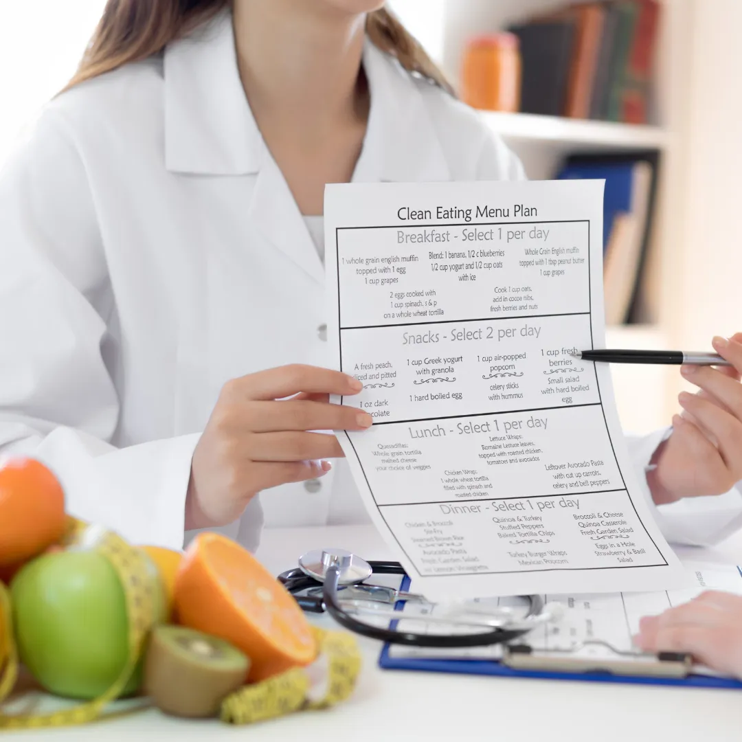 Nutritionist or healthcare professional in a white coat holding a printed clean eating menu plan while discussing it with a patient. A clipboard, stethoscope, and a variety of fresh fruits are visible on the desk, emphasizing a focus on healthy eating and dietary guidance.