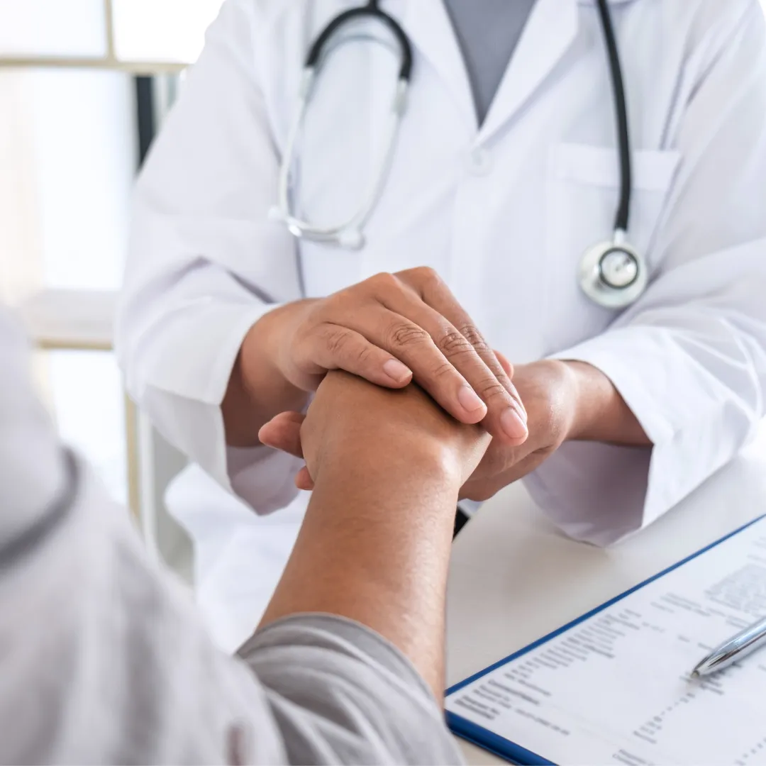 Close-up of a doctor in a white coat offering comfort by gently holding a patient’s hands. A stethoscope hangs around the doctor’s neck, and a medical form with a pen is visible on the desk, indicating a supportive consultation or discussion.