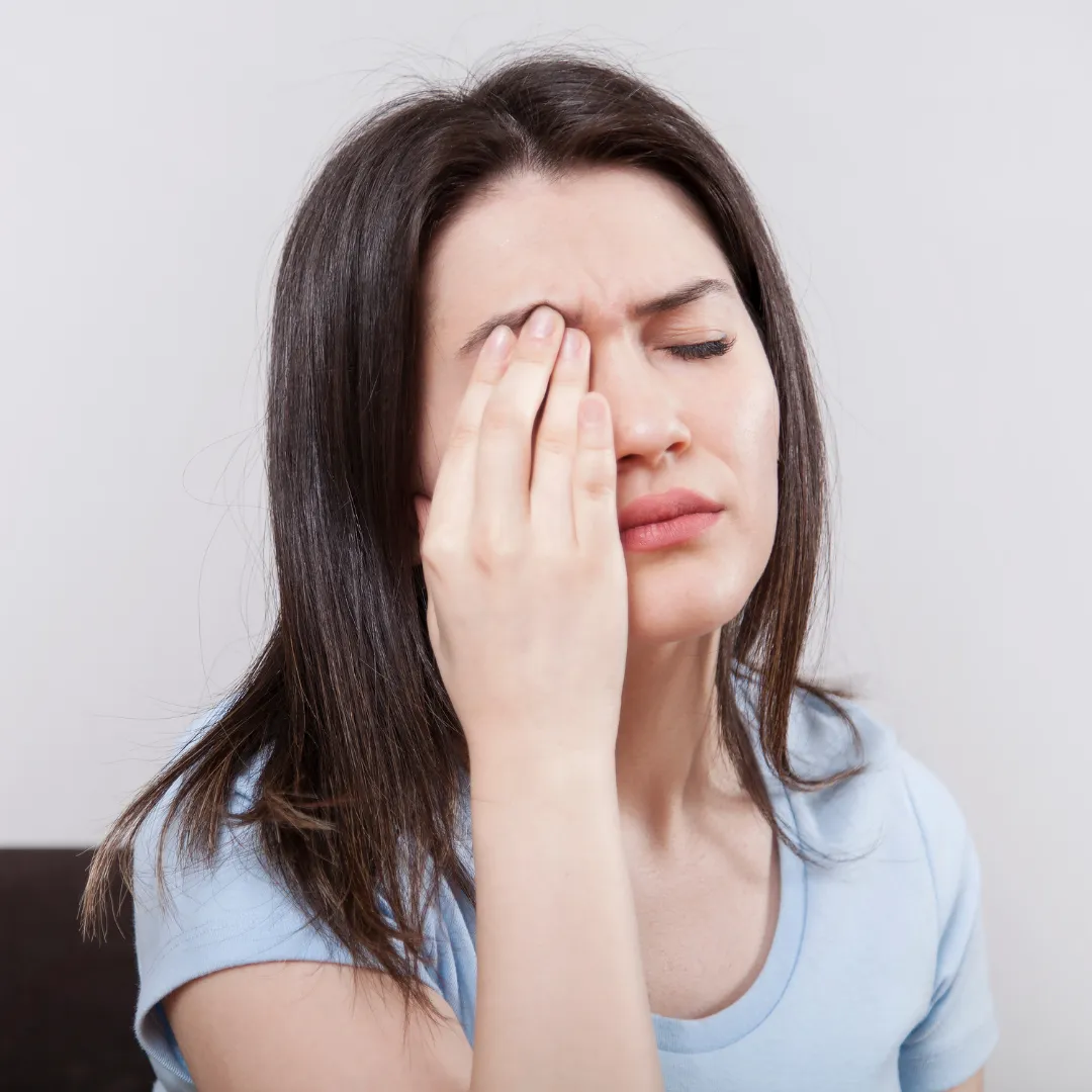 Young woman in a light blue shirt holding her hand to her forehead and closing her eyes with a pained expression, indicating discomfort or a headache.