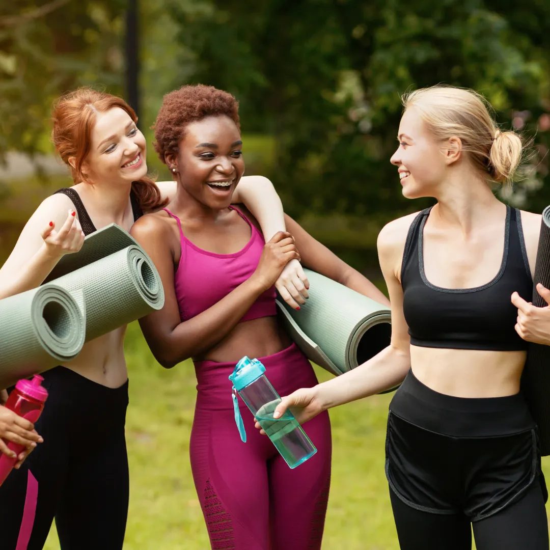 Three smiling women in activewear holding yoga mats and water bottles, standing outdoors in a park. They appear to be chatting and enjoying each other’s company after a fitness or yoga session.