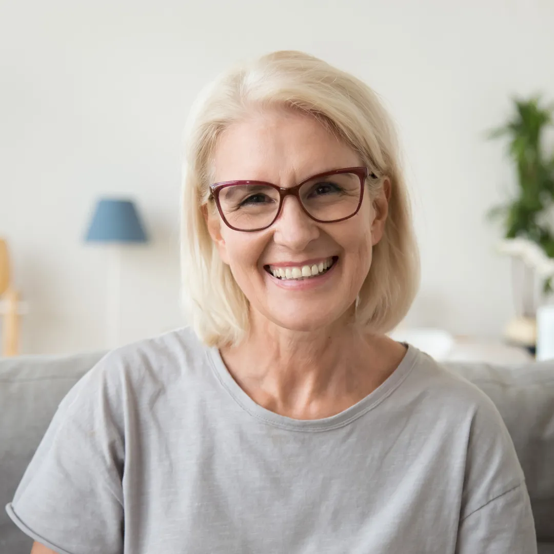 Smiling older woman with short blonde hair and glasses, wearing a light grey shirt. She is seated indoors on a sofa in a cozy, well-lit living room with a soft background featuring a lamp and plants.