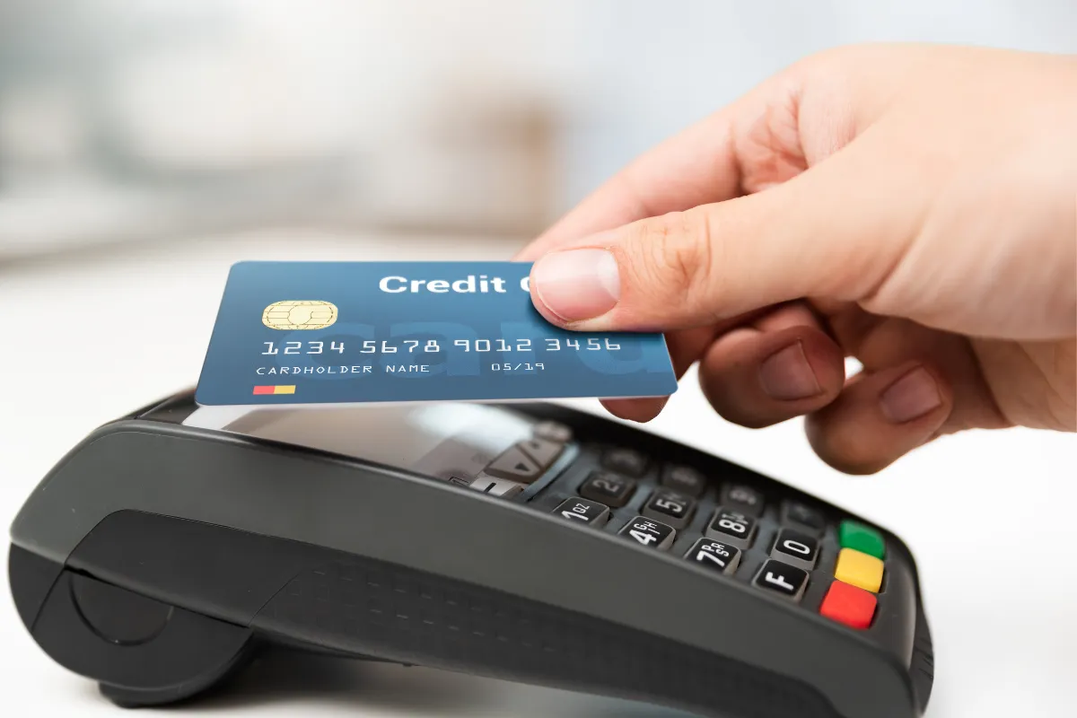 Close-up of a hand holding a blue credit card above a payment terminal, preparing to tap for a contactless transaction. The scene suggests a retail or point-of-sale environment.