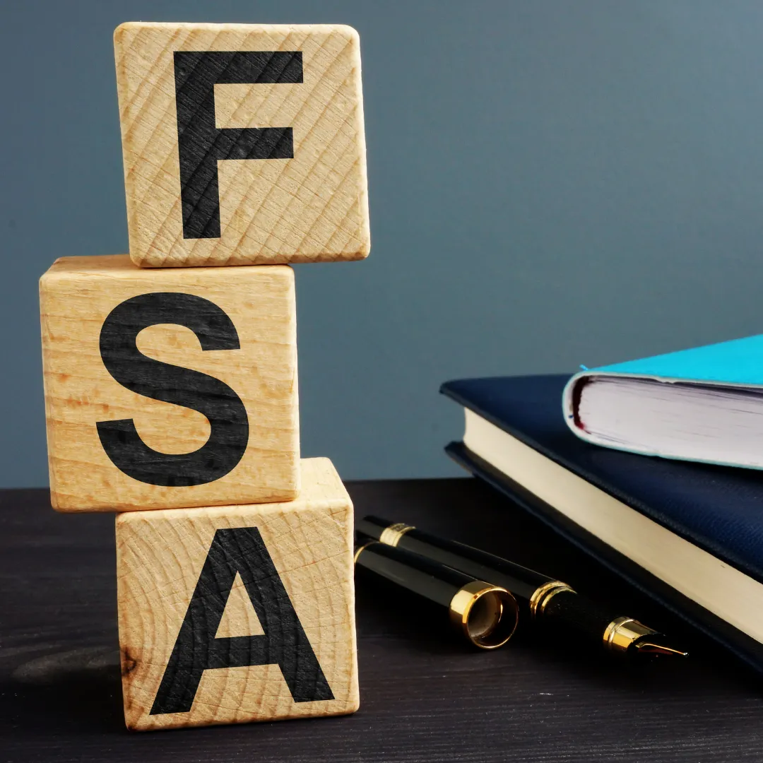 Three wooden blocks stacked vertically with large black letters spelling ‘FSA.’ The blocks are placed on a dark surface next to a fountain pen and a closed notebook, representing the concept of a Flexible Spending Account.