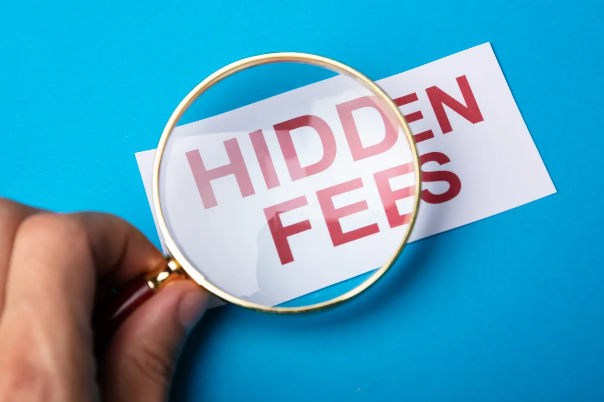 A hand holding a magnifying glass over a white paper with the words ‘HIDDEN FEES’ in bold red letters, placed on a blue background. The magnifier emphasizes the text, symbolizing scrutiny of concealed charges.