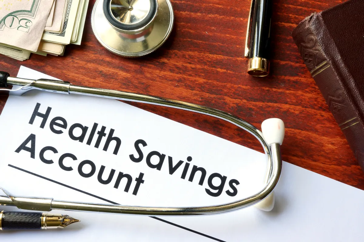 Document labeled ‘Health Savings Account’ on a wooden desk surrounded by a stethoscope, cash, fountain pen, and a leather-bound book, representing financial planning for healthcare.