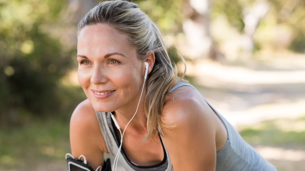 Active woman with earphones taking a break from exercising outdoors. She is smiling slightly, wearing a grey tank top, and appears energised and relaxed in a sunlit natural setting.