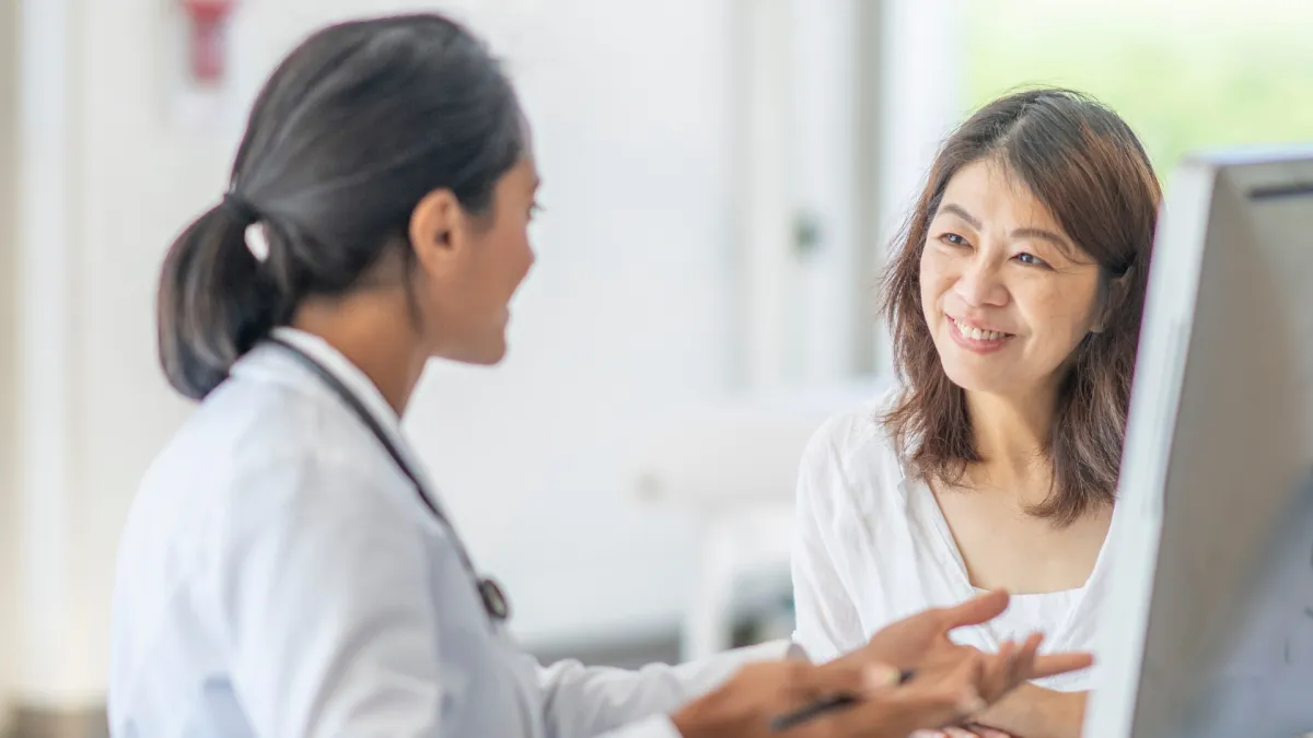Female doctor speaking with a smiling female patient in a bright, modern medical office. The doctor is gesturing with her hands while the patient listens attentively, indicating a positive and supportive consultation.