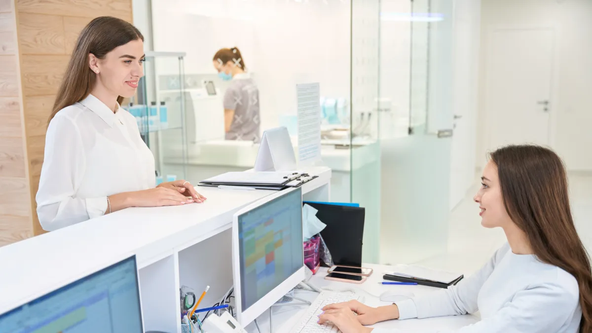 Receptionist at a medical or dental clinic desk speaking with a smiling female patient. Two computer monitors are on the counter, and a staff member in scrubs and a face mask can be seen working in the background through a glass wall.