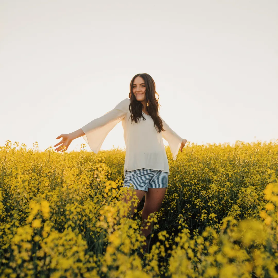 Young woman smiling with arms outstretched while standing in a vibrant yellow flower field under a clear sky. She is wearing a light blouse and striped shorts, enjoying the peaceful outdoor setting.