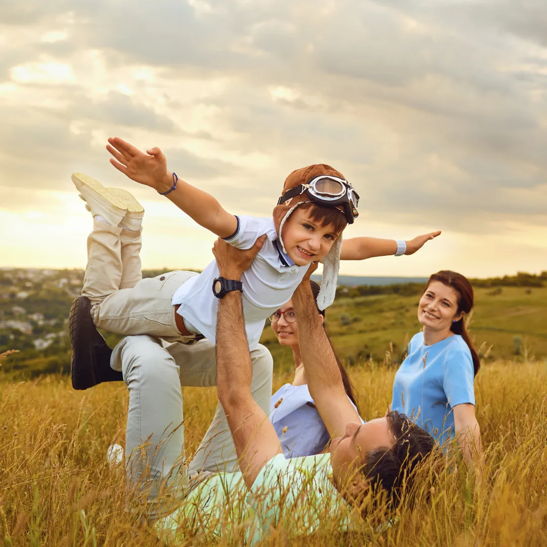 Happy family playing together in a grassy field at sunset. A child wearing aviator goggles is being lifted into the air by an adult, pretending to fly, while two other family members smile and watch. The atmosphere is joyful and carefree.
